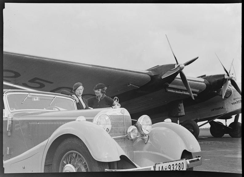 Two women, a Mercedes-Benz 500K convertible car, and aeroplane