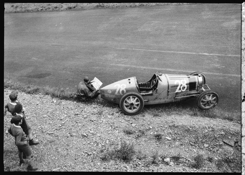 A driver reads a newspaper beside his Bugatti racing car