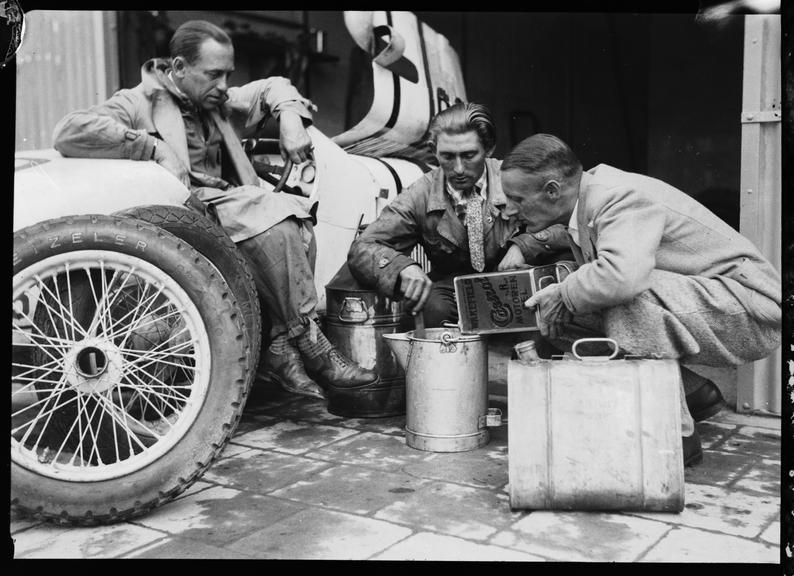 Gerhard Macher in DKW racing car while mechanics mix oil and