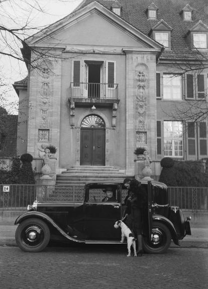 Woman with dog enters black Mercedes Benz, in front of house