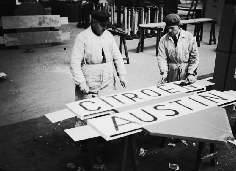 Two men in a workshop with 'Citroen' and 'Austin' signs