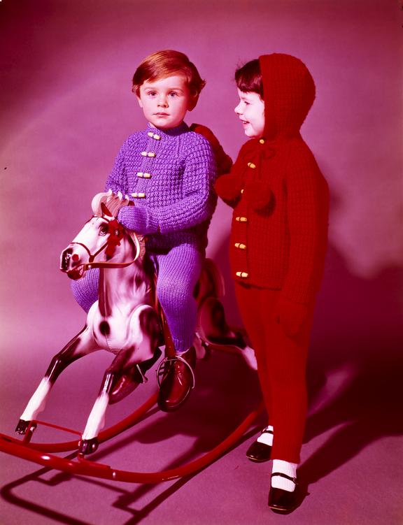 Girl in knitted outfit stands next to boy on rocking horse