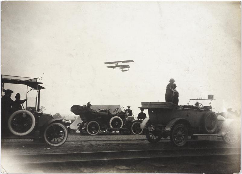 Photograph of a byplane flying over some cars