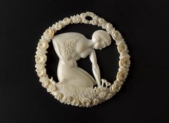 Carved pendant of imitation ivory. Shows woman picking roses, surrounded by wreath of roses