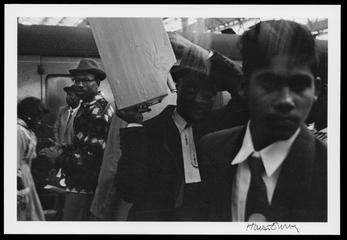 Man carrying a box on his shoulder amongst a group of people on a platform next to a railway carriage
